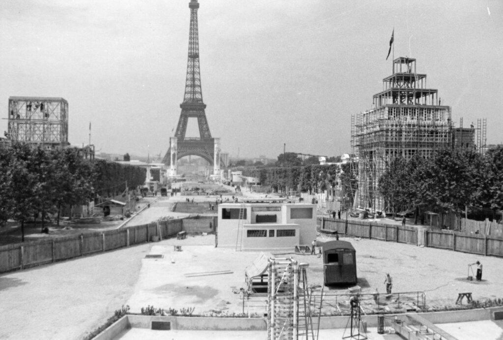 Photographie noir et blanc de l'installation d'une cabine de projection sur le parvis du palais de la lumière et de l'électricité. La Tour Eiffel apparait en arrière plan.